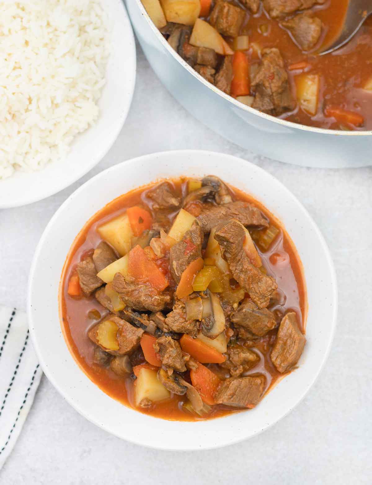A bowl of beef stew with tomato sauce and a bowl of rice is in the background.