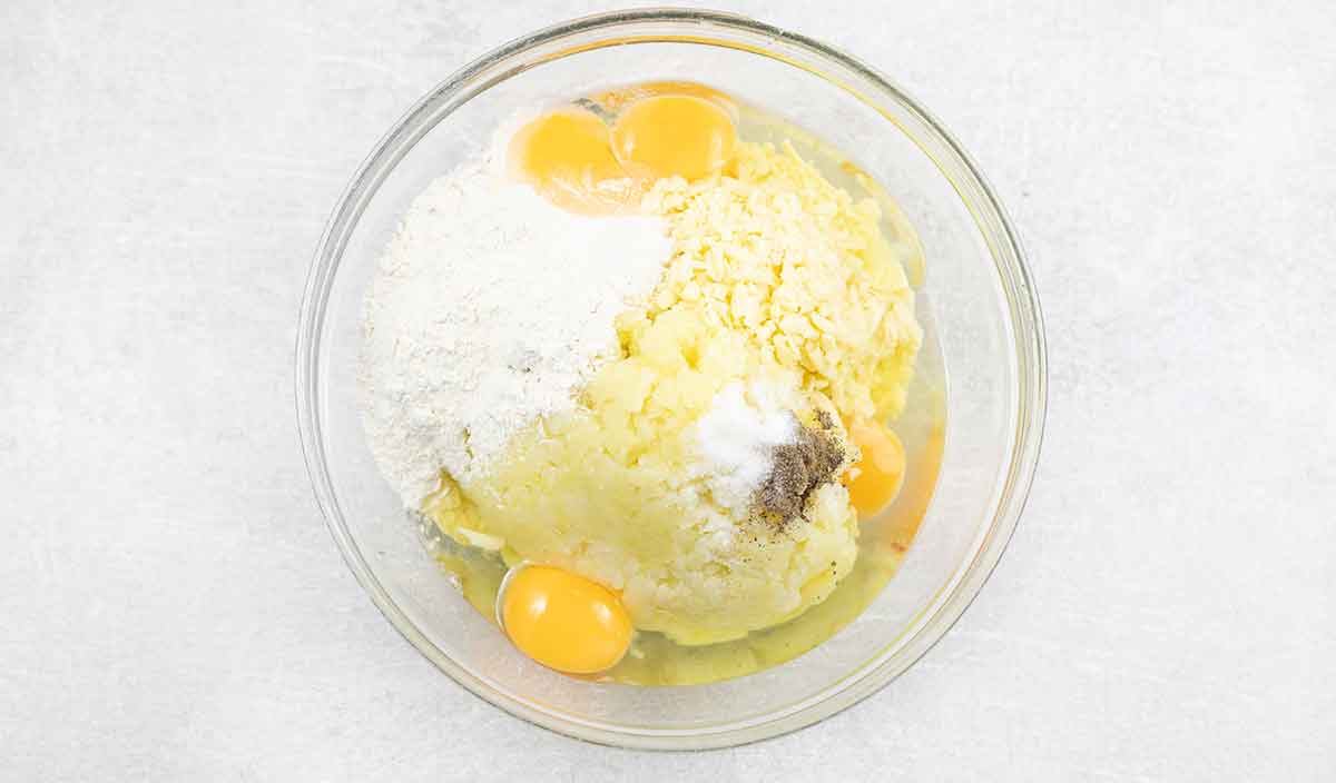 mashed potatoes, flour, eggs and cheese are in a mixing bowl.